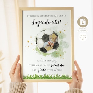 Op de afbeelding: Een ingelijste print met een voetbalontwerp versierd met eurobiljetten. De print bevat Duitse tekst, gouden sterren en groen gras aan de onderkant. De print wordt door een persoon vastgehouden.