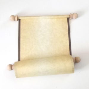 May include: A parchment-style scroll featuring a textured, light beige paper surface with dark brown vertical borders. The scroll is attached to two smooth, light-colored wooden rods at the top and bottom, each finished with rounded wooden finials.