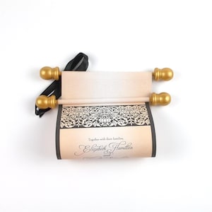 May include: A rolled invitation with gold-colored finials and a black ribbon. The invitation features a decorative black and white border and the text "Together with their families, Elizabeth Hamilton and..."