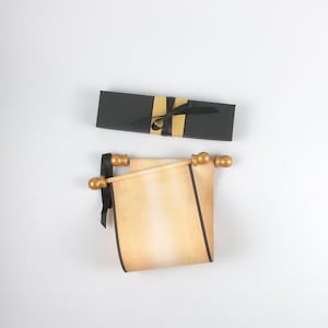 May include: A beige parchment scroll with dark edges and gold-coloured finials on the ends, accompanied by a black rectangular gift box wrapped with a gold band and a black ribbon. The scroll is rolled and tied with a black ribbon on one side.