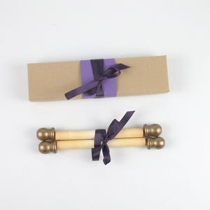 May include: Two scroll invitations with gold-colored finials and a dark purple ribbon. A kraft paper box is also shown, wrapped with a purple band and a dark purple ribbon. The items are on a white background.