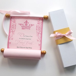 May include: Pink and gold scroll with a crown and the text "Once Upon a Time in a land far away lived Princess Emily Queen Kasey and King Phillip". The scroll is rolled up and tied with a pink ribbon. It is in a white box with a gold ribbon.