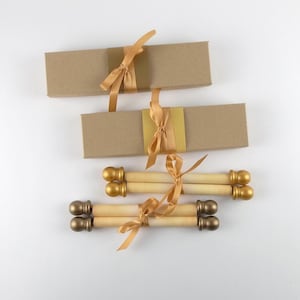May include: Two rectangular brown cardboard boxes wrapped with gold satin ribbons and two rolled paper scrolls with gold-colored finials at each end, each tied with a matching gold satin ribbon, arranged on a plain white surface.
