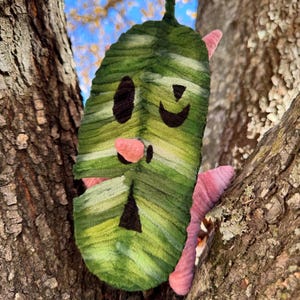 May include: A whimsical, handmade felt leaf with a smiling face, nestled in a tree. The leaf is primarily green with lighter green and yellow accents, and features black eyes and a smiling mouth. Pink accents are visible on the sides.