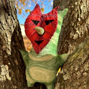 May include: A whimsical, handmade plush creature with a red face, black facial features, and a green body. The creature is nestled in the branches of a tree, with a blue sky and fall foliage in the background. The plush has a unique, textured appearance.