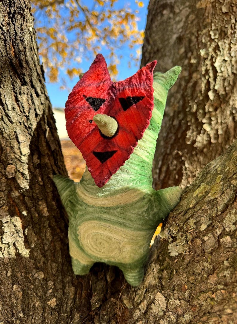 May include: A whimsical, handmade plush creature with a red face, black facial features, and a green body. The creature is nestled in the branches of a tree, with a blue sky and fall foliage in the background. The plush has a unique, textured appearance.