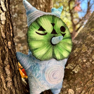 May include: A whimsical plush creature with a green watermelon-like face and a blue and white body. The plush has a cone-shaped nose and a winking eye. It is perched in a tree, with the bark visible in the background.