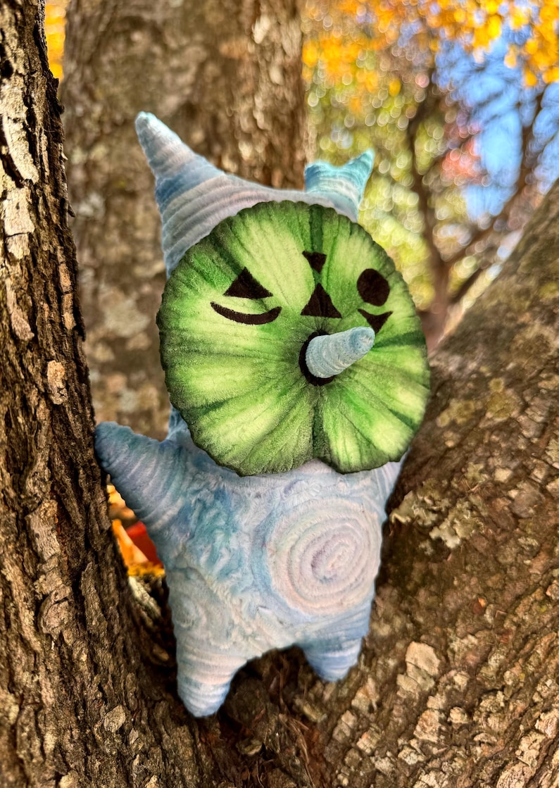 May include: A whimsical plush creature with a green watermelon-like face and a blue and white body. The plush has a cone-shaped nose and a winking eye. It is perched in a tree, with the bark visible in the background.
