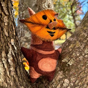 May include: A handmade felt creature with an orange leaf-shaped face, a small brown nose, and a smiling black mouth. The body is brown with lighter brown accents and a circular design. The plush toy is positioned in a tree.