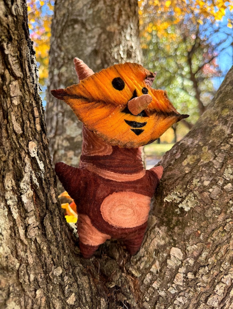 May include: A handmade felt creature with an orange leaf-shaped face, a small brown nose, and a smiling black mouth. The body is brown with lighter brown accents and a circular design. The plush toy is positioned in a tree.