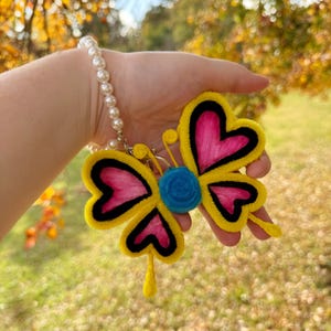 Peut inclure: Un porte-clés en forme de papillon jaune avec des ailes roses et un centre bleu. Le papillon a des contours noirs et est fait de feutre.