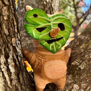 May include: A plush Korok doll, featuring a leaf-shaped face with embroidered details and a small, cone-shaped nose. The doll's body is brown with a textured pattern, and it is perched in a tree.