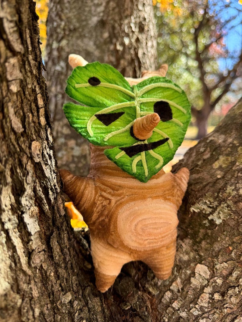 May include: A plush Korok doll, featuring a leaf-shaped face with embroidered details and a small, cone-shaped nose. The doll's body is brown with a textured pattern, and it is perched in a tree.