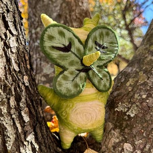 May include: A handmade, whimsical plush creature with a green and yellow color scheme, resembling a stylized insect or mythical being. It has leaf-like wings and is nestled in a tree, creating a charming, nature-inspired aesthetic.