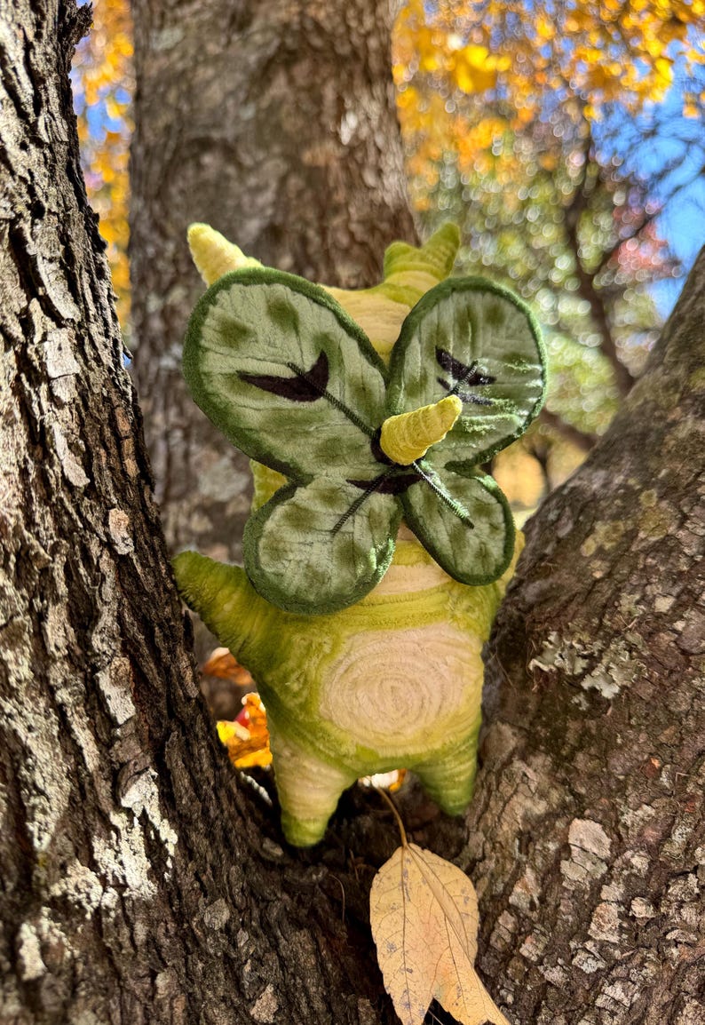 May include: A handmade, whimsical plush creature with a green and yellow color scheme, resembling a stylized insect or mythical being. It has leaf-like wings and is nestled in a tree, creating a charming, nature-inspired aesthetic.