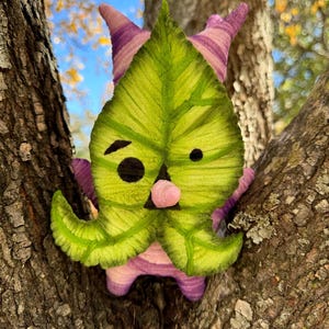 May include: A whimsical, handmade plush toy shaped like a leaf with a face, perched in a tree. The leaf is green with detailed veining, and the toy has pink and purple accents, including pointed ears and a small nose.