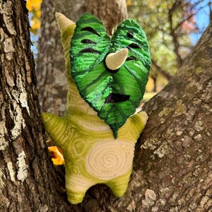 May include: A handmade plush toy in the shape of a Korok character, featuring a green leaf-shaped face with black details and a white horn. The body is a light green with a spiral design. The toy is set against a tree trunk.
