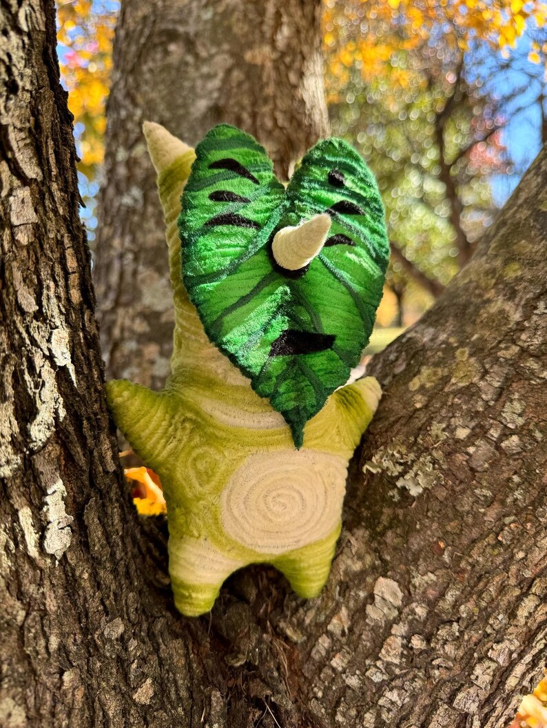 May include: A handmade plush toy in the shape of a Korok character, featuring a green leaf-shaped face with black details and a white horn. The body is a light green with a spiral design. The toy is set against a tree trunk.