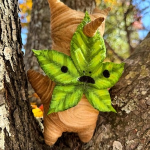 May include: A handmade plush figure of a creature with a leaf face and a brown, textured body. The green leaf face has black dots for eyes and a small black mouth. The figure is perched on a tree branch.