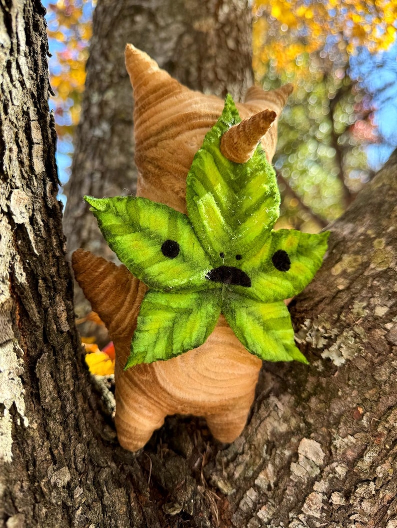 May include: A handmade plush figure of a creature with a leaf face and a brown, textured body. The green leaf face has black dots for eyes and a small black mouth. The figure is perched on a tree branch.