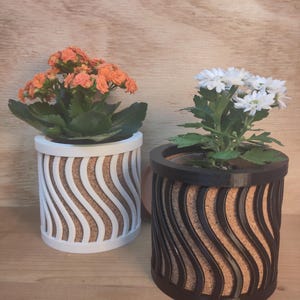 Könnte beinhalten: Zwei dekorative Blumentöpfe mit Blumen. Einer ist weiß mit einem wellenförmigen Design und orangefarbenen Blumen. Der andere ist schwarz mit einem wellenförmigen Design und weißen Blumen. Beide Töpfe haben ein korkartiges Material in der Mitte.