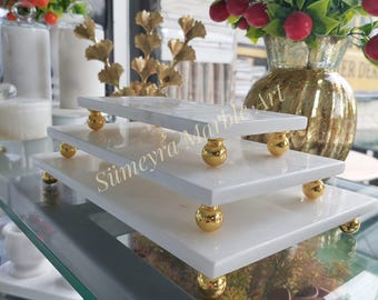 以下が含まれることがあります： 金色のアクセントが付いた白い大理石のサービングトレイが3つ。トレイは積み重ねられており、各コーナーに装飾的な金色のボールが付いています。トレイはガラスの表面に置かれています。トレイには「Siimeyra Marble Art」という文字が見えます。