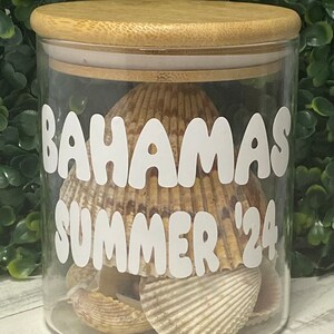 May include: A clear glass jar with a bamboo lid filled with seashells. The jar has a white vinyl sticker with the text "Bahamas Summer '24" in a playful font.