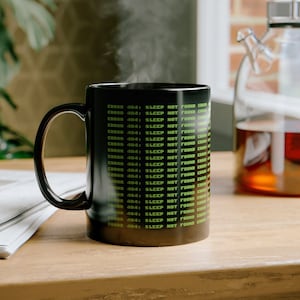 May include: A black coffee mug with a green and white design that reads "ERROR 404: SLEEP NOT FOUND". The mug is sitting on a wooden table with a stack of newspapers and a glass teapot in the background.