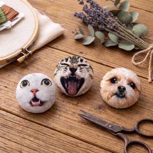 Puede incluir: Tres cojines redondos de felpa con caras de gato y perro con la boca abierta, sobre una superficie de madera. Tambi&eacute;n hay un par de tijeras antiguas y un manojo de lavanda. Un bastidor de bordado y carretes de hilo est&aacute;n en el fondo.