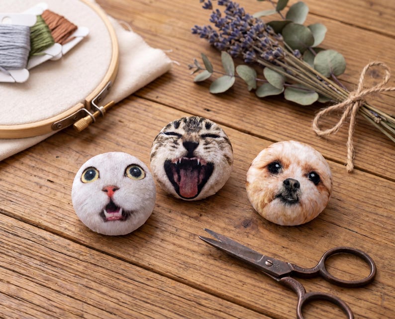 Puede incluir: Tres cojines redondos de felpa con caras de gato y perro con la boca abierta, sobre una superficie de madera. Tambi&eacute;n hay un par de tijeras antiguas y un manojo de lavanda. Un bastidor de bordado y carretes de hilo est&aacute;n en el fondo.