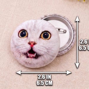 Puede incluir: Un bot&oacute;n redondo con la imagen de la cara de un gato sorprendido. El bot&oacute;n mide 6,5 cm de di&aacute;metro y tiene una parte trasera de metal con un imperdible. El gato tiene grandes ojos amarillos y pelaje blanco y gris.