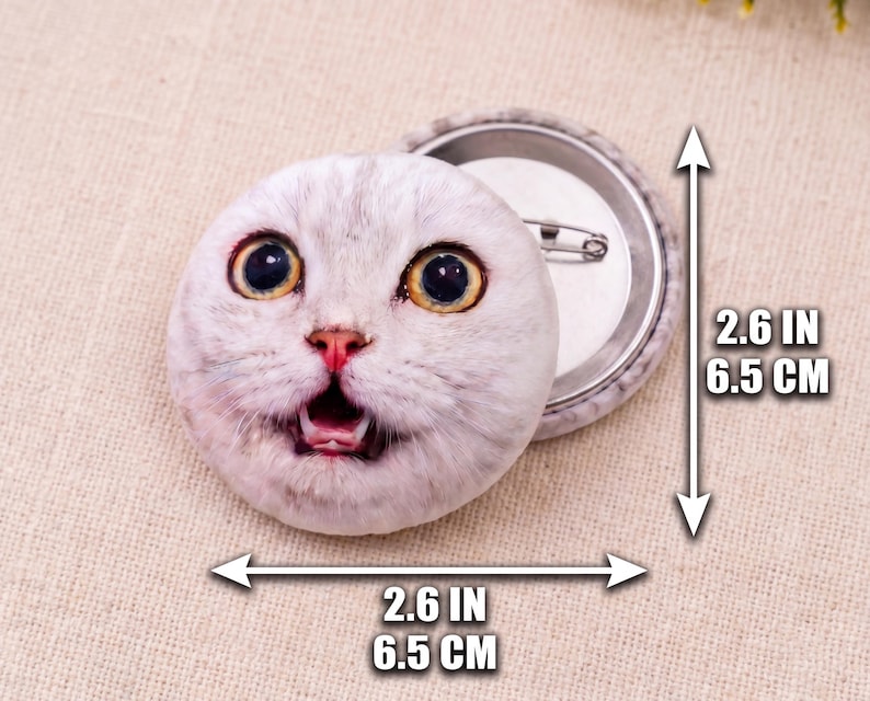 Puede incluir: Un bot&oacute;n redondo con la imagen de la cara de un gato sorprendido. El bot&oacute;n mide 6,5 cm de di&aacute;metro y tiene una parte trasera de metal con un imperdible. El gato tiene grandes ojos amarillos y pelaje blanco y gris.