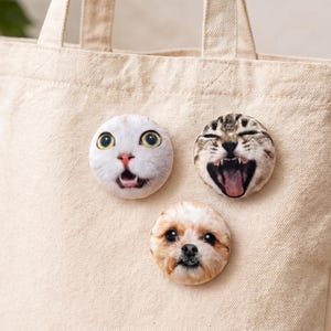Puede incluir: Tres imanes redondos con caras de animales est&aacute;n adheridos a una bolsa tote beige. Los imanes presentan un gato blanco con ojos grandes, un gato atigrado con la boca abierta y un perro peque&ntilde;o con pelaje marr&oacute;n y blanco.