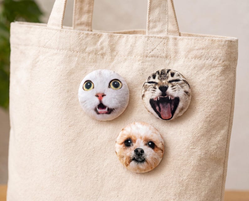 Puede incluir: Tres imanes redondos con caras de animales est&aacute;n adheridos a una bolsa tote beige. Los imanes presentan un gato blanco con ojos grandes, un gato atigrado con la boca abierta y un perro peque&ntilde;o con pelaje marr&oacute;n y blanco.