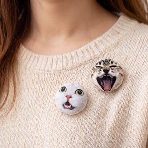 Puede incluir: Dos broches redondos con caras de gato. Uno muestra una cara de gato blanco con la boca abierta, el otro una cara de gato atigrado con la boca abierta y la lengua fuera. Los broches est&aacute;n sujetos a un su&eacute;ter beige.