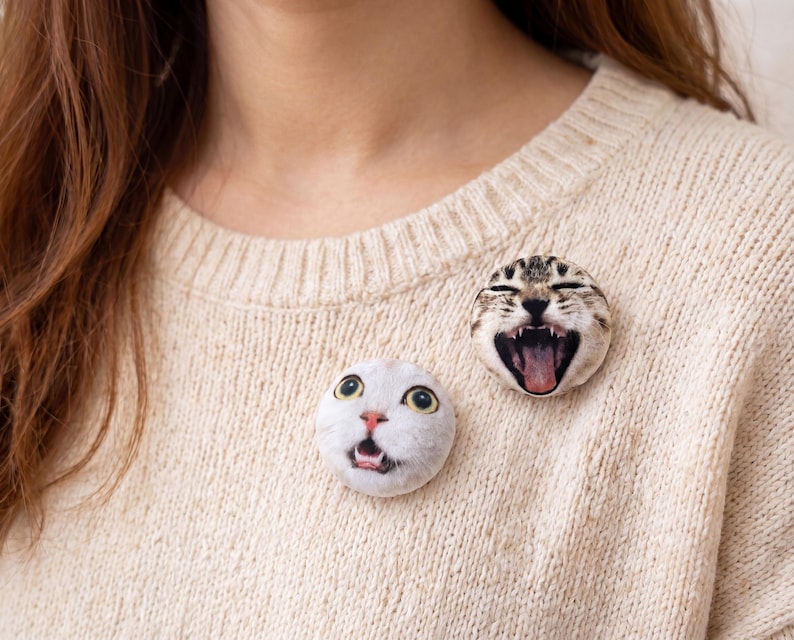 Puede incluir: Dos broches redondos con caras de gato. Uno muestra una cara de gato blanco con la boca abierta, el otro una cara de gato atigrado con la boca abierta y la lengua fuera. Los broches est&aacute;n sujetos a un su&eacute;ter beige.