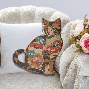 May include: A wooden cat-shaped cushion with a colourful vintage design. The cushion features a cat with green eyes and a red, white and blue pattern. The cushion is sitting on a white sofa with a white cushion behind it.