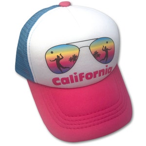 May include: White trucker hat with a pink bill. The front of the hat features a pair of sunglasses with a beach scene reflected in the lenses. The text "California" is printed below the sunglasses in pink.