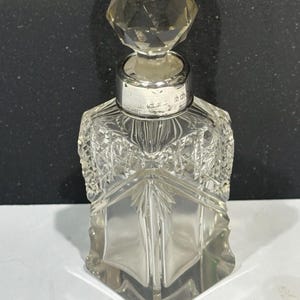 Può includere: Un decanter in vetro trasparente con base quadrata e tappo geometrico sfaccettato. Il collo è decorato con un collare in argento con marchi incisi. Il corpo del decanter presenta un dettagliato motivo a taglio incrociato nella parte superiore.