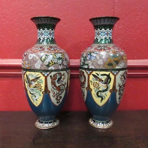Peut inclure: Une paire de vases cloisonnés assortis présentant un travail d'émail complexe. Le motif comprend des dragons détaillés, des fleurs et des bordures géométriques dans des tons de bleu, vert, jaune et blanc. Chaque vase a un col et une base évasés.