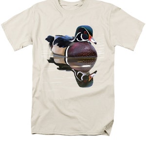 May include: A beige t-shirt with a colorful illustration of a wood duck swimming in water. The duck has a green head, red eyes, a white stripe, and a brown body with white spots. The duck's reflection is visible in the water.