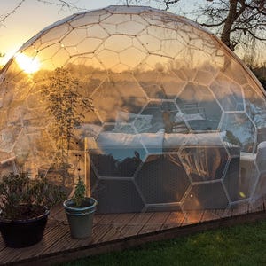 May include: A clear, geodesic dome structure with a wooden deck base, set in a grassy outdoor space. The dome's interior features seating, and potted plants are placed on the deck. The setting sun casts a warm glow through the transparent panels.