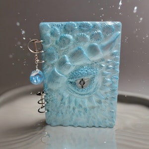 May include: A light blue notebook with a dragon eye design and a silver spiral binding. A silver charm with a light blue glass orb hangs from the binding.