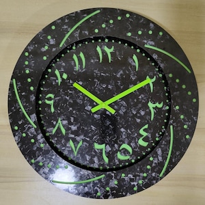 Op de afbeelding: Een ronde wandklok met een zwart gespikkelde wijzerplaat en neon groene Arabische cijfers en wijzers. De klok heeft een zwarte buitenring met groene accenten en een reeks groene stippen. Het ontwerp is modern en opvallend.
