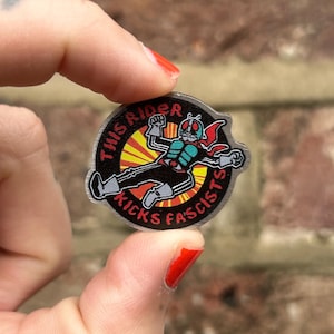 May include: A circular badge with a cartoon figure in a red cape and helmet, and the text "THIS RIDER KICKS FASCISTS". The design features a yellow and red burst behind the character, set on a silver background.