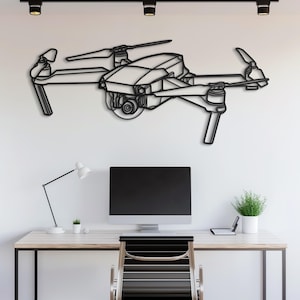 Op de afbeelding: Zwarte metalen drone-wandkunst. De drone heeft gedetailleerde contouren van de behuizing, propellers en camera. Het kunstwerk is bevestigd aan een witte muur boven een bureau met een computermonitor, bureaulamp en een kleine plant.