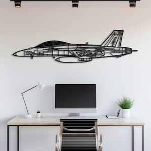 Pode incluir: Uma peça de arte de parede em metal preto que retrata um contorno detalhado de um caça. A obra de arte é montada em uma parede branca acima de uma mesa com um monitor de computador e material de escritório. O design do jato inclui detalhes internos intrincados.