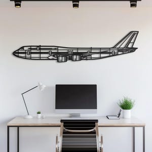 以下が含まれることがあります： ボーイング747航空機の輪郭を描いた黒い金属製の壁掛けアート。アートワークは、コンピューターモニター、デスクランプ、小さな鉢植えの植物を備えたデスクの上にある白い壁に取り付けられています。飛行機の内部構造が見えます。
