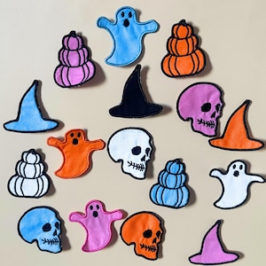 May include: A collection of embroidered patches featuring Halloween-themed designs including ghosts, skulls, pumpkins, and witch hats in various colours such as blue, orange, pink, white, and black, arranged in a scattered pattern on a light background.