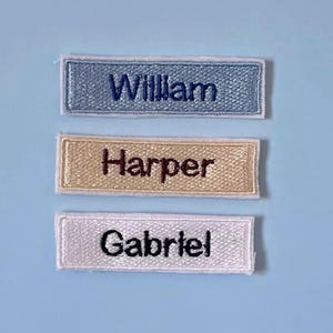 May include: Three rectangular embroidered name patches with white borders. The top patch is light blue with dark blue text reading William. The middle patch is beige with dark brown text reading Harper. The bottom patch is white with black text reading Gabriel.